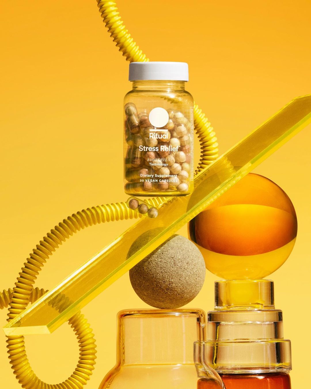 Still life shot of a Ritual Stress Relief supplement bottle surrounded by glass orbs and shapes on a yellow background. Product photography.