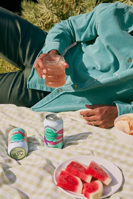 Person lying on a picnic blanket with cans of beverages, a glass of a pink drink, slices of watermelon, and snack outdoors.