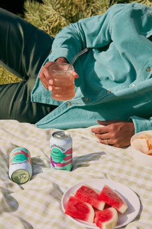 Person lying on a picnic blanket with cans of beverages, a glass of a pink drink, slices of watermelon, and snack outdoors.