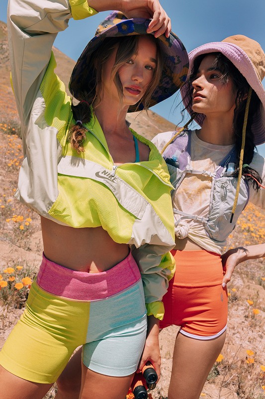 Two women in athletic wear and bucket hats stand outdoors in a sunny, hilly landscape. They wear shorts, jackets, and hiking gear.