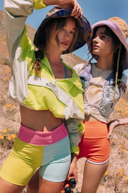 Two women in athletic wear and bucket hats stand outdoors in a sunny, hilly landscape. They wear shorts, jackets, and hiking gear.
