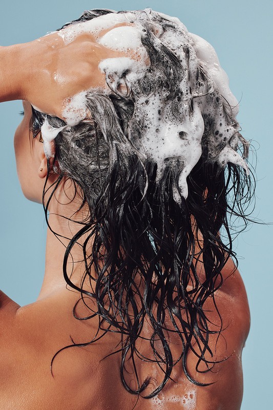 Back view of a brunette woman washing her hair with shampoo. A hand is seen applying the shampoo to her hair against light blue background.