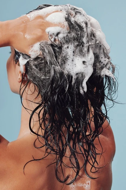 Back view of a brunette woman washing her hair with shampoo. A hand is seen applying the shampoo to her hair against light blue background.