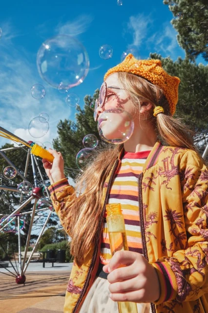 Young girl blows bubbles at the park on a sunny day. She wears an orange hat, round glasses, and a patterned jacket. Blue sky, trees.