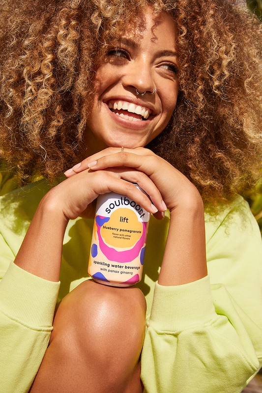 Smiling woman with curly hair holds a can of Soulboost Lift sparkling water beverage with ginseng while wearing a green sweatshirt outdoors.