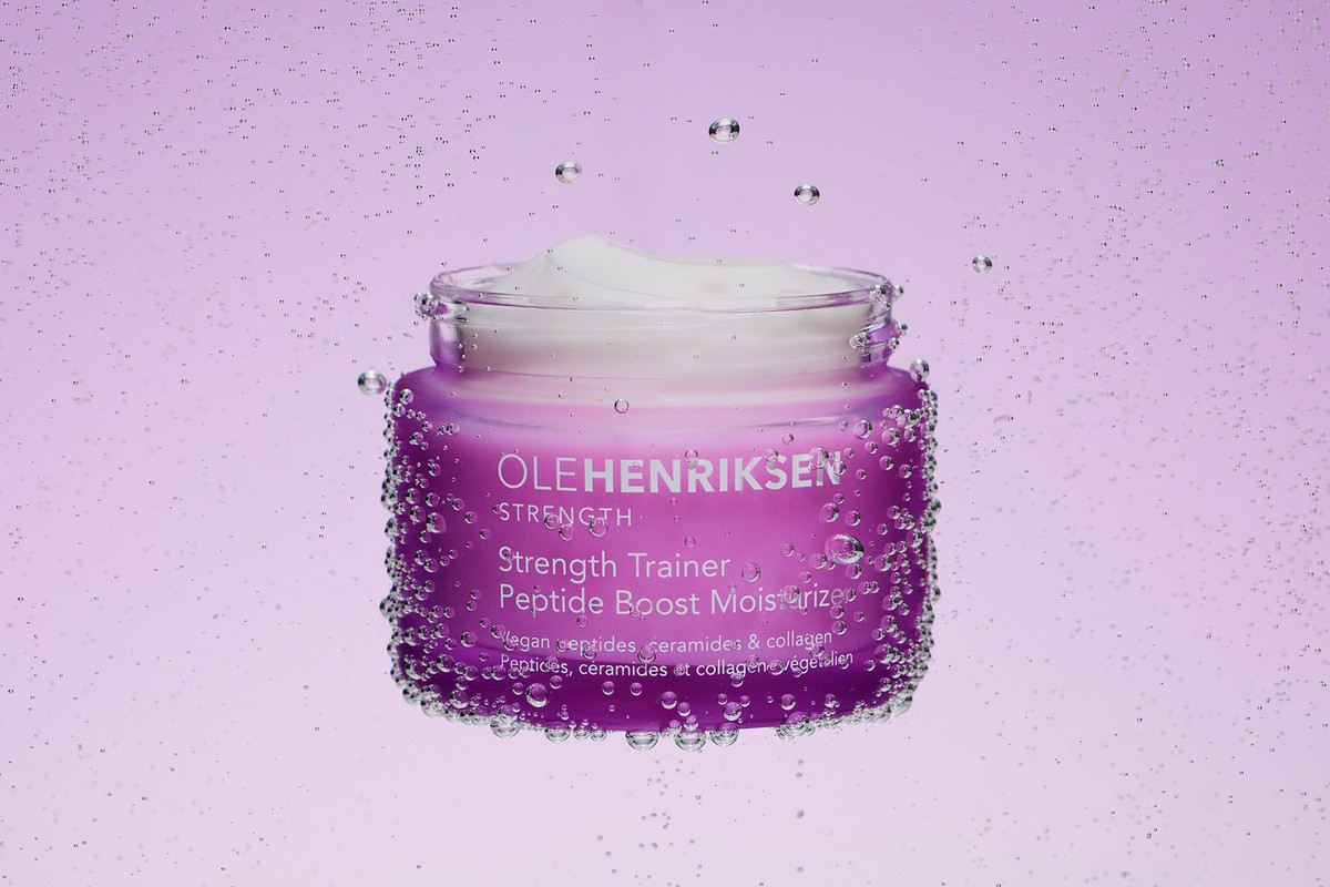 Ole Henriksen skincare product, Strength Trainer Peptide Boost Moisturizer, in a purple jar with water droplets against a purple gradient.