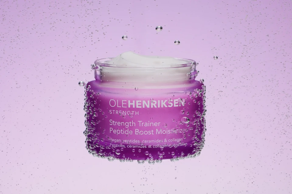 Ole Henriksen skincare product, Strength Trainer Peptide Boost Moisturizer, in a purple jar with water droplets against a purple gradient.