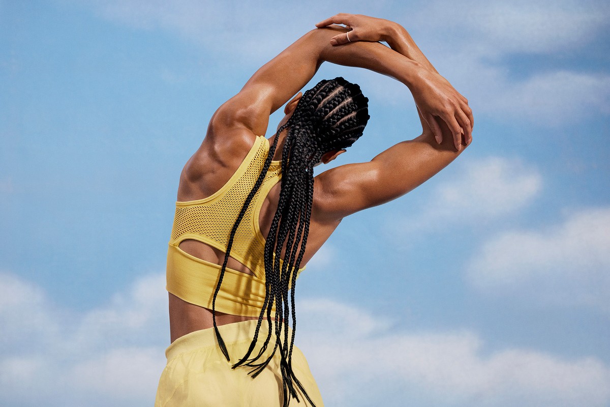 Woman in yellow sports bra stretches arms overhead against blue sky. Athletic fitness workout outdoors with active lifestyle in sunshine.