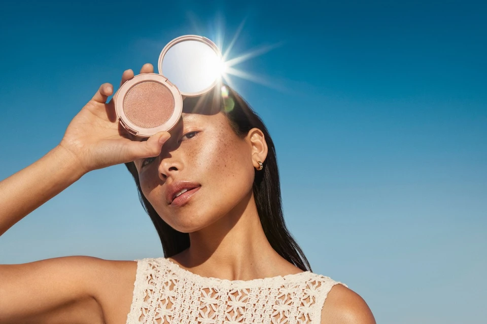 Sunlit portrait of a young Asian woman with freckles holding a round compact makeup case against a clear blue sky, beauty shot.