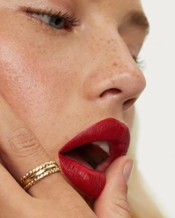 Close-up portrait of a woman with red lipstick, freckles, and stacked gold rings on her finger against a neutral white background