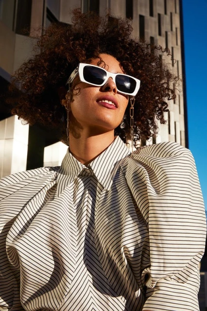 A woman with curly hair and sunglasses wears a striped shirt and poses outdoors with a modern building in the background.