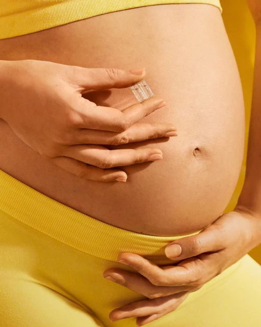 Closeup of pregnant woman holding transparent object against her abdomen, wearing yellow top and pants in a brightly lit setting.