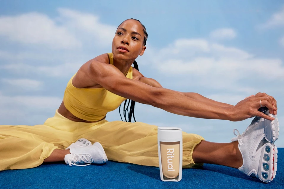 Athletic woman in yellow activewear stretching outdoors with a fitness drink, set against a blue sky with soft white clouds.