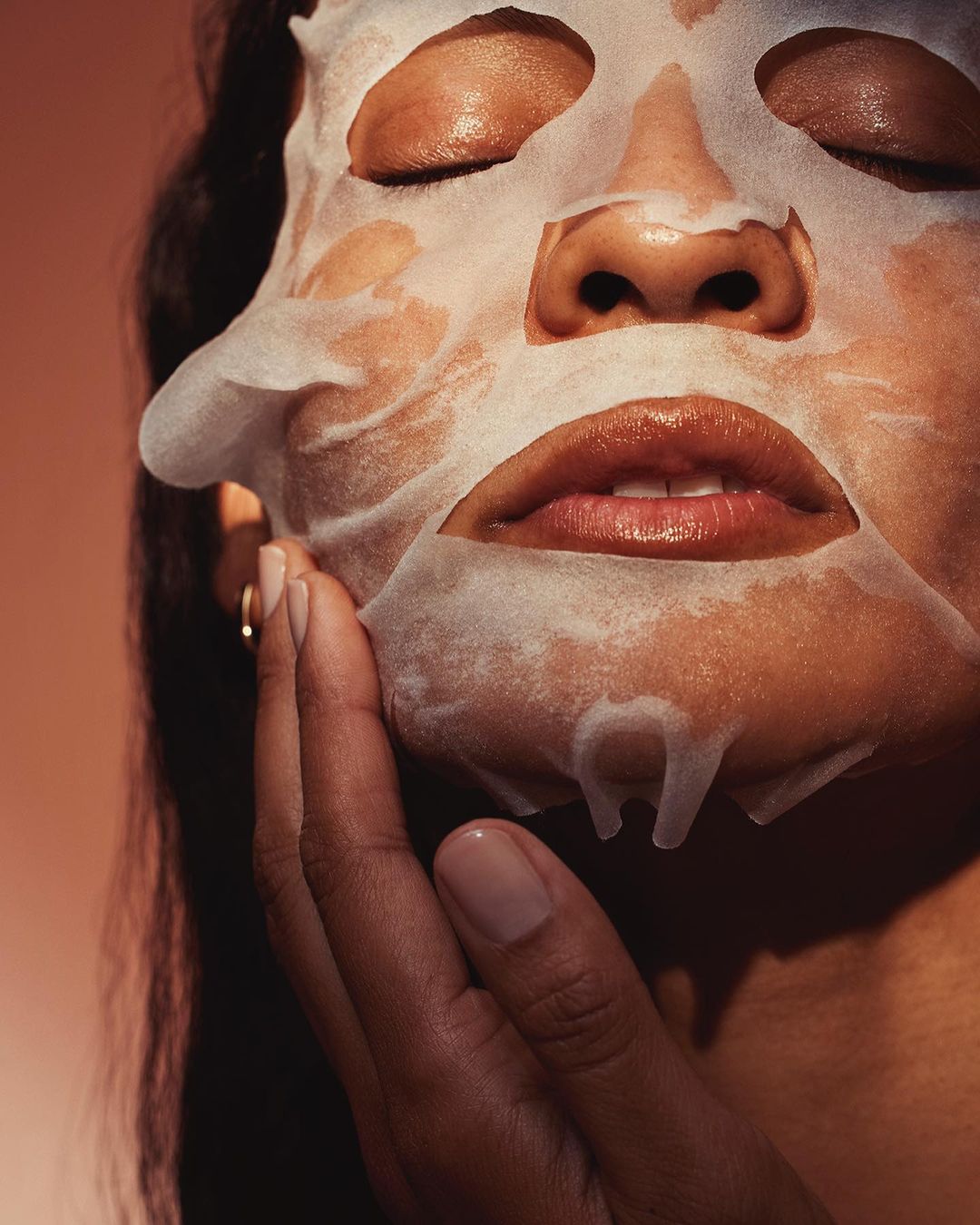 Close-up of an adult female with a sheet mask on her face, eyes closed, touching her face with her hand against a peach gradient background.