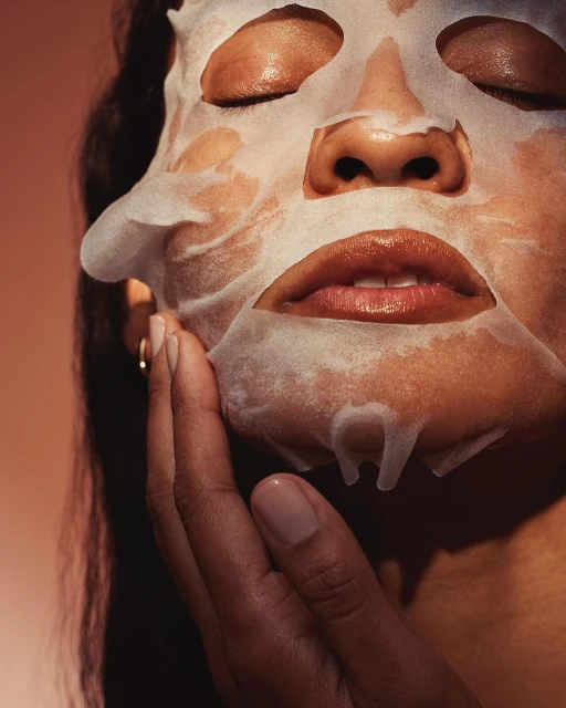 Close-up of an adult female with a sheet mask on her face, eyes closed, touching her face with her hand against a peach gradient background.