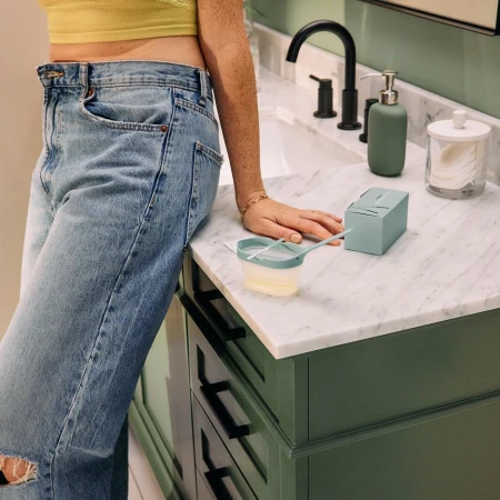 A woman stands in a bathroom next to a green vanity with a white marble top and a collection of self-care items.