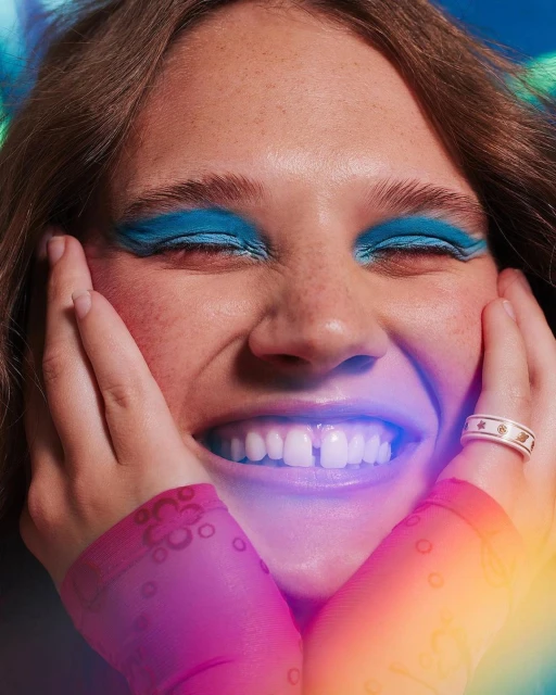 Close-up of smiling woman with bold blue eyeshadow, hands on cheeks, wearing ring, fair skin, brown hair, colorful gradient background.