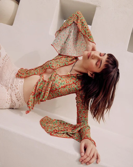 A woman in a floral top poses on a white surface, eyes closed, with her arm raised above her head and a necklace around her neck.