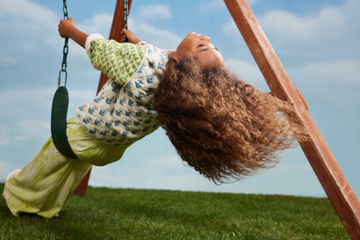 Child swings on a playground swing set with curly brown hair, wearing a printed jacket and yellow pants, with a blue sky background.
