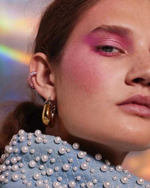 Close-up of a young woman with bold pink eye makeup, hoop earrings, and a pearl-studded denim jacket in front of a holographic background.