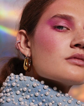 Close-up of a young woman with bold pink eye makeup, hoop earrings, and a pearl-studded denim jacket in front of a holographic background.