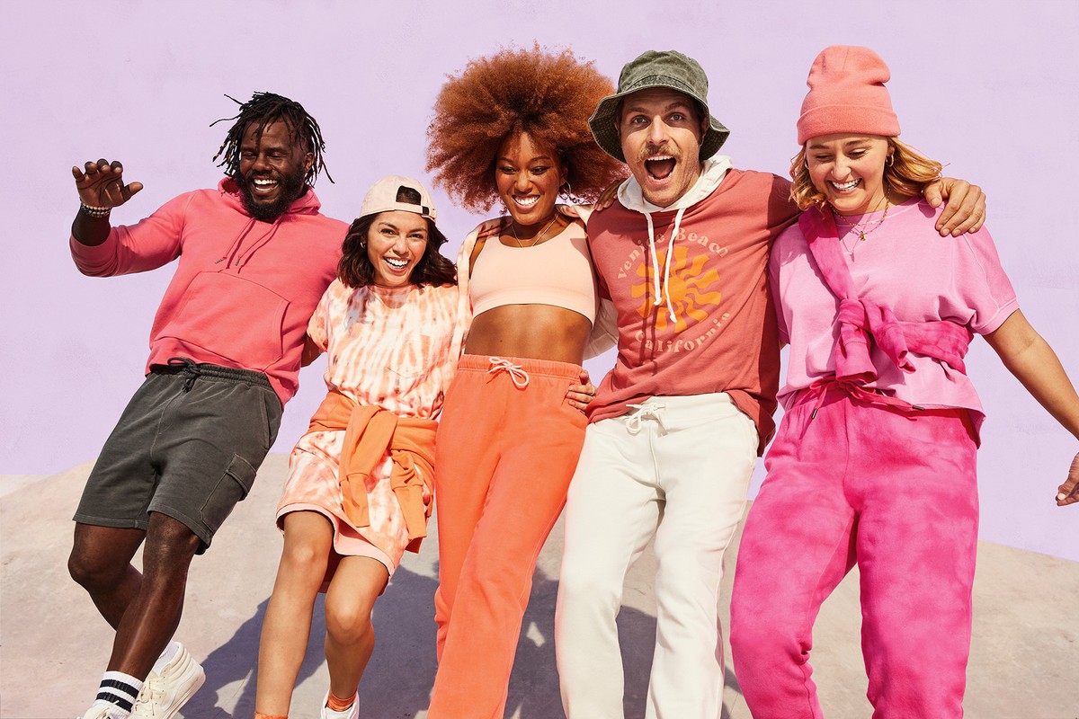 Five diverse people, smiling and posing in bright casual clothing against a lilac background, showcasing a fun, colorful fashion collection.