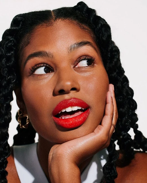 Close-up portrait of a young Black woman with twists looking to the side, wearing bright red lipstick, gold earrings, and a white top.