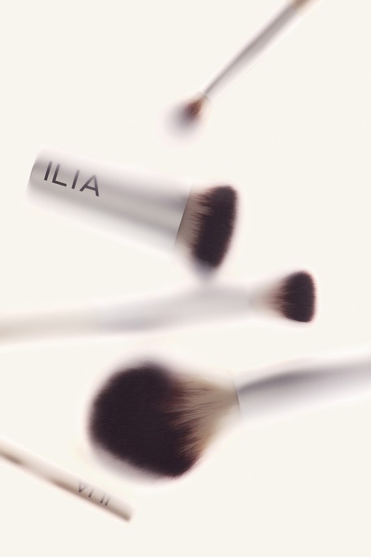 Several Ilia makeup brushes with white handles and dark bristles are suspended mid-air against a soft, neutral background.