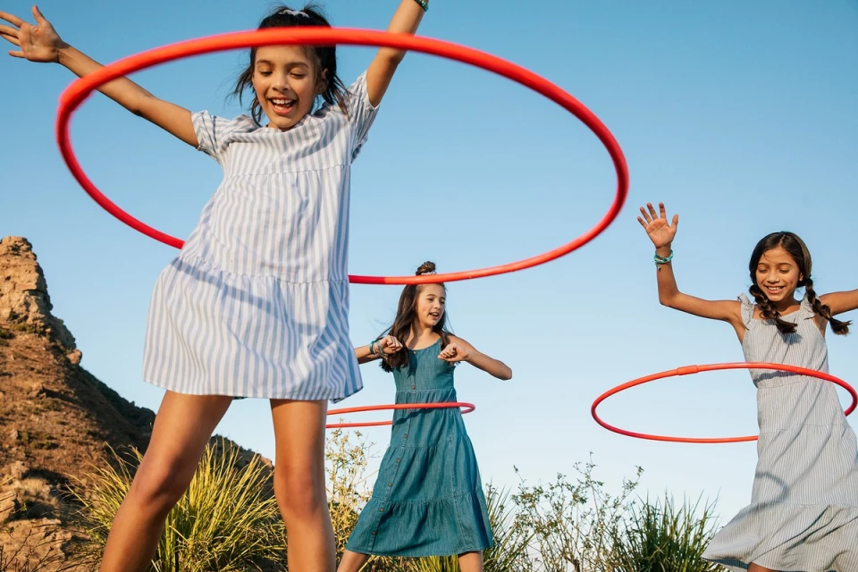 Three young girls are playing outdoors with red hula hoops against a clear blue sky, having fun in the sunshine with a rocky hill.