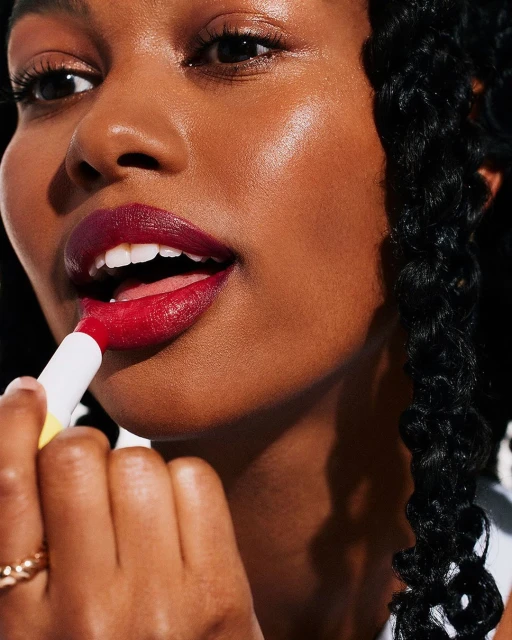 Close-up of a woman with dark skin applying red lipstick with a white and yellow applicator. The background is out of focus.