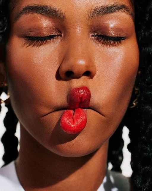 Close-up of a woman with curly hair, closed eyes, and shiny skin, her red lips puckered in a playful expression.