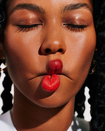 Close-up of a woman with curly hair, closed eyes, and shiny skin, her red lips puckered in a playful expression.