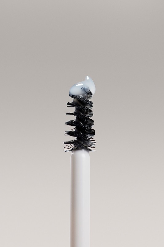 Close-up of a mascara wand applicator brush with a small amount of white cosmetic product on the bristles against a solid backdrop.