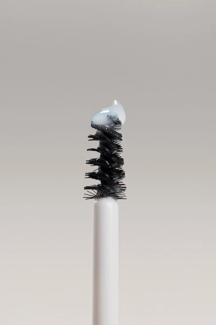 Close-up of a mascara wand applicator brush with a small amount of white cosmetic product on the bristles against a solid backdrop.