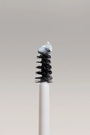 Close-up of a mascara wand applicator brush with a small amount of white cosmetic product on the bristles against a solid backdrop.