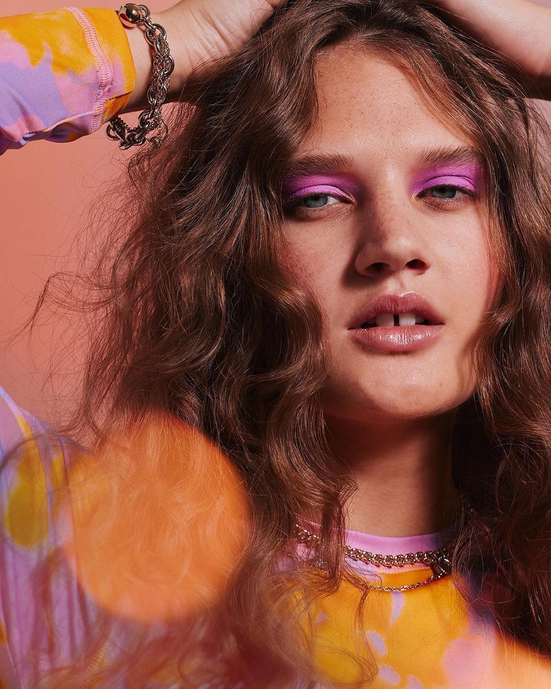 Portrait of a young woman with curly hair and pink eyeshadow. She poses with her hands in her hair against a peach background.