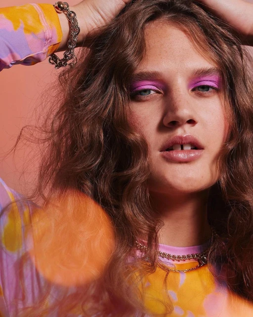 Portrait of a young woman with curly hair and pink eyeshadow. She poses with her hands in her hair against a peach background.