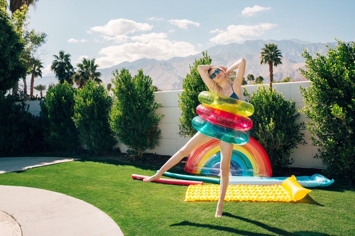 Woman in swim suit poses with rainbow and colorful inflatable rings in a sunny backyard with green grass and mountain views.