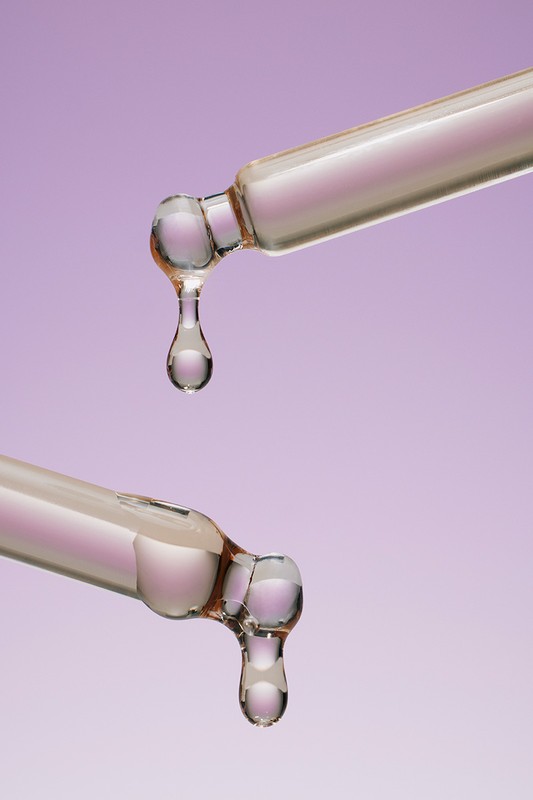 Close-up of two glass droppers with translucent liquid droplets against a light purple backdrop, showcasing beauty or pharmaceutical product.