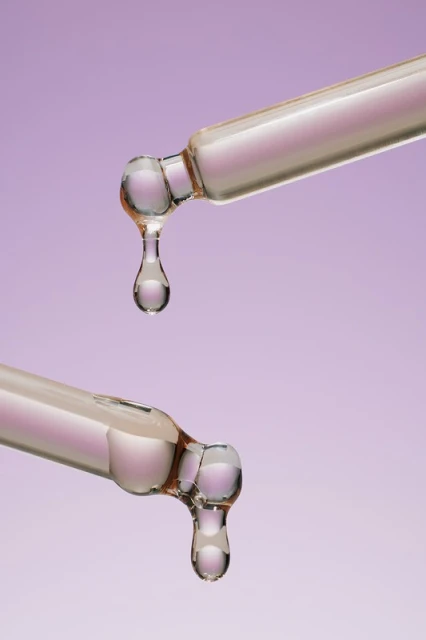 Close-up of two glass droppers with translucent liquid droplets against a light purple backdrop, showcasing beauty or pharmaceutical product.