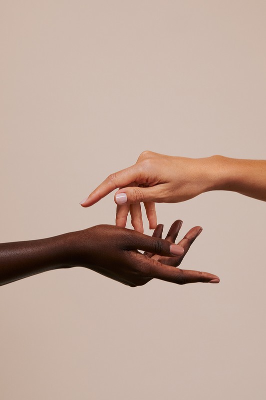 Two hands reach toward each other against a neutral background. One hand is fair-skinned, the other is dark-skinned. Close-up studio shot.