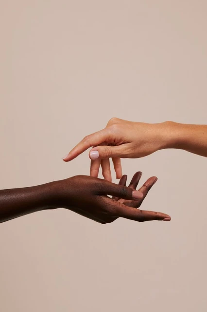 Two hands reach toward each other against a neutral background. One hand is fair-skinned, the other is dark-skinned. Close-up studio shot.