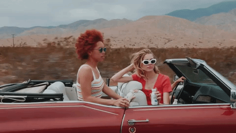 Two women enjoy a road trip in a red convertible car, driving through a desert landscape with mountains in the background.