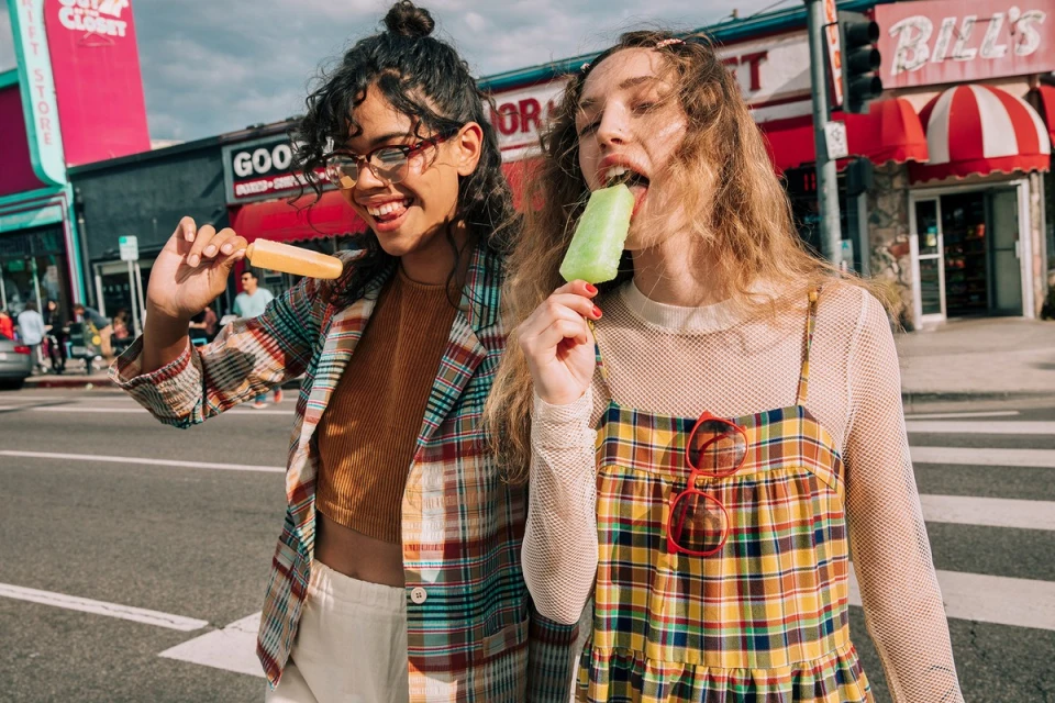 Two women stroll outdoors eating popsicles, showcasing warm casual style in a city setting with buildings and street details under a bright sky.