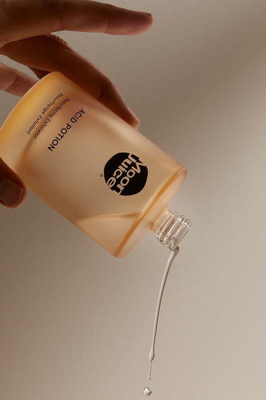 Aesthetic product shot of a person holding an open bottle of Moon Juice Acid Potion toner with liquid pouring out of it against a plain backdrop.
