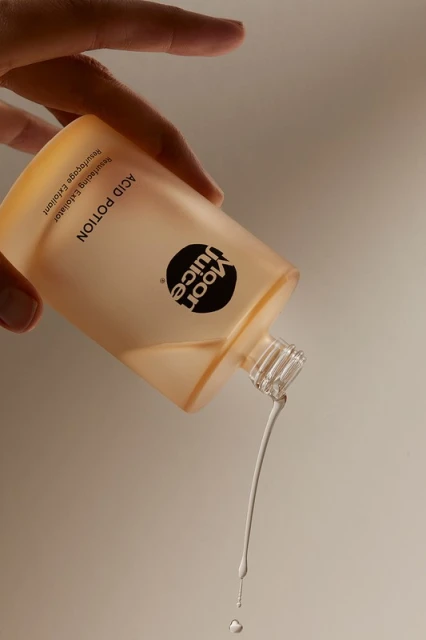Aesthetic product shot of a person holding an open bottle of Moon Juice Acid Potion toner with liquid pouring out of it against a plain backdrop.