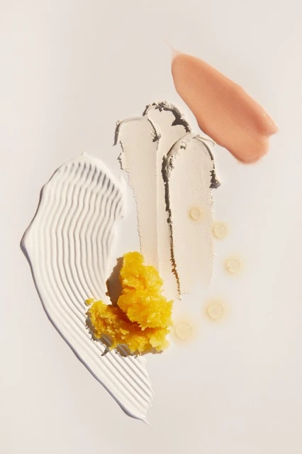 Overhead shot of skincare product smears in white, beige, and yellow on a white surface, creating a textured beauty product display.