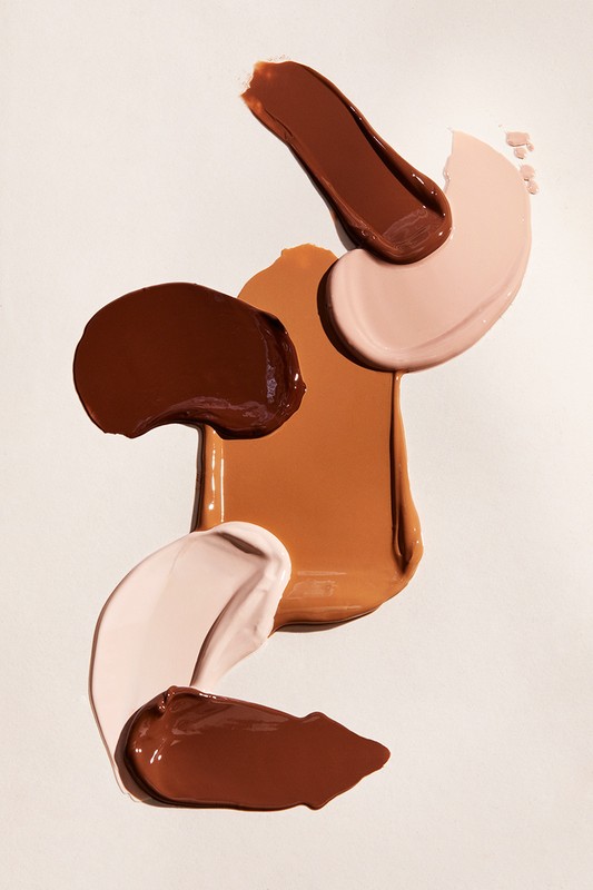 Close-up of cosmetic foundation swatches in different shades of brown and beige, floating on a white background. Beauty product shot.
