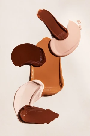 Close-up of cosmetic foundation swatches in different shades of brown and beige, floating on a white background. Beauty product shot.