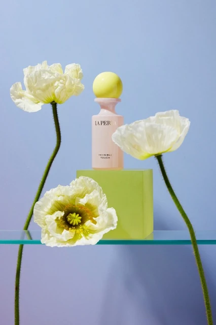 Still life of a pink perfume bottle with a yellow cap on a neon green block, with white poppy flowers on a light blue background.